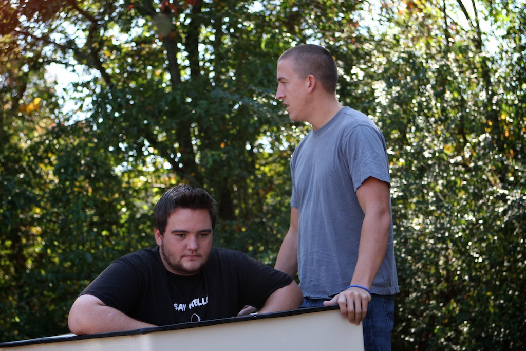 two men standing behind a large white object near trees