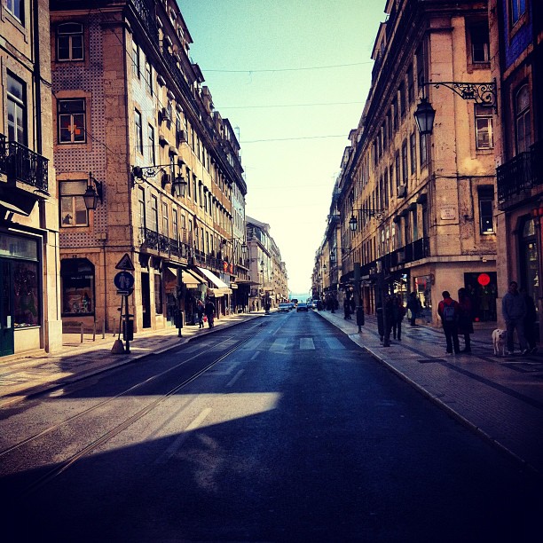 a street in the middle of a city, with people walking and a yellow traffic light at the end