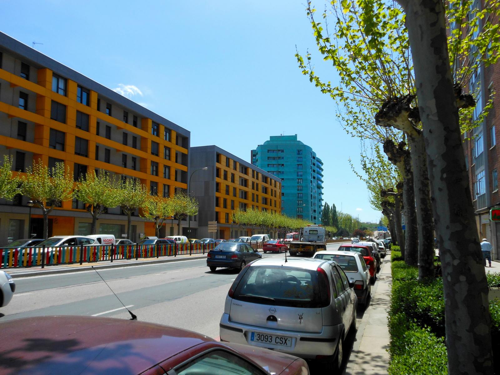a busy street that has parked cars along one side and multiple colorful buildings behind the cars