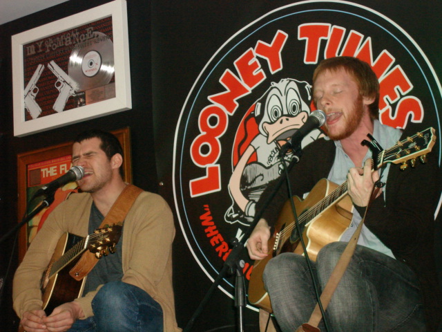 two men are on stage with one holding an acoustic guitar