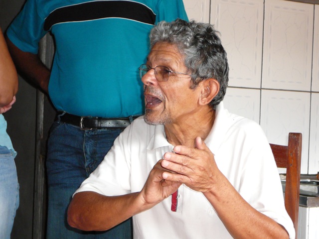 an older man in a room sitting at a table talking to someone