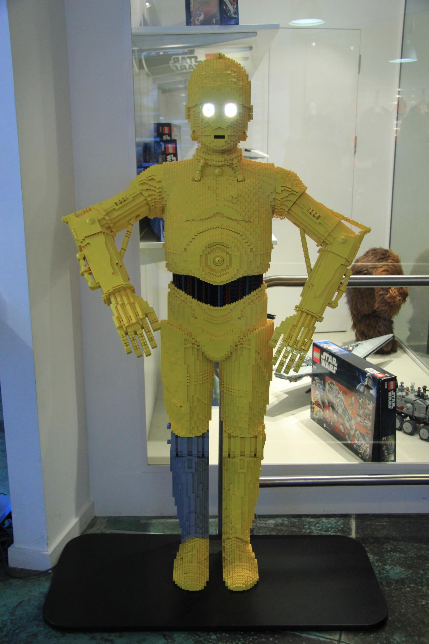 a robot made with yellow plastic legos sits in front of a glass display