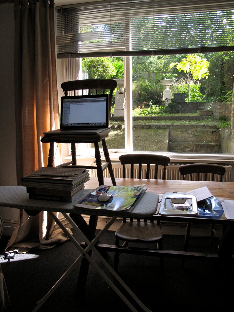 the large computer on top of the wooden table is next to a window