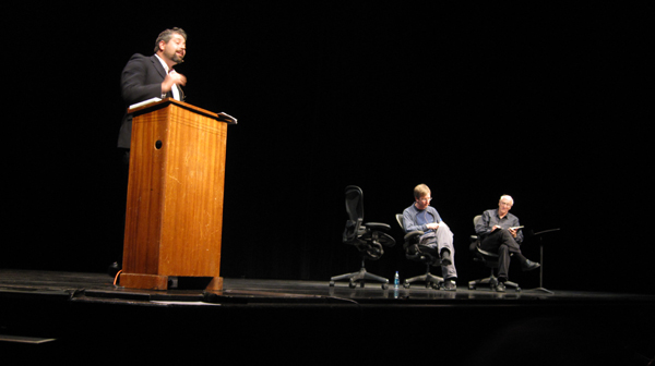 three people are sitting at a podium while a man stands
