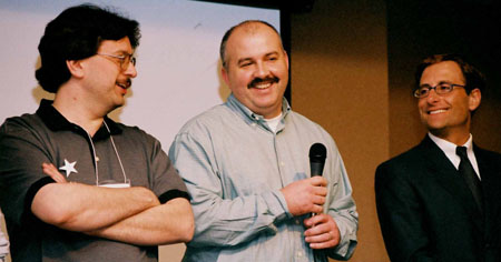 three men are laughing and talking while a man with glasses stands up