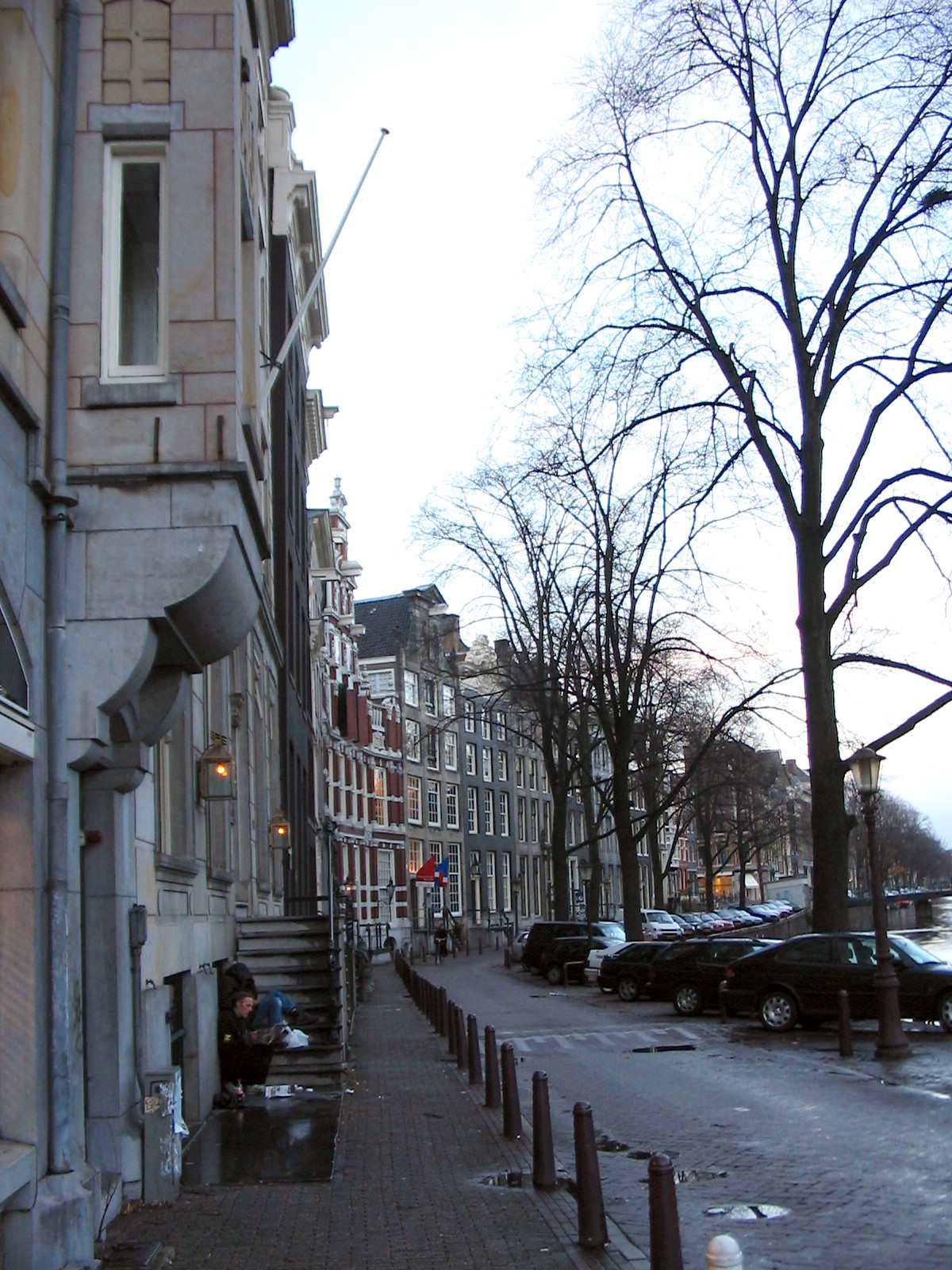 a city street with buildings, trees, and a man on a bench