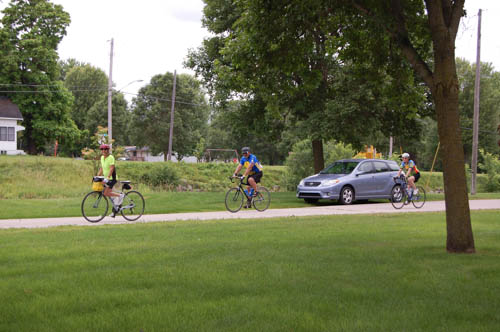 people are riding bikes on a road and two cars parked in the background