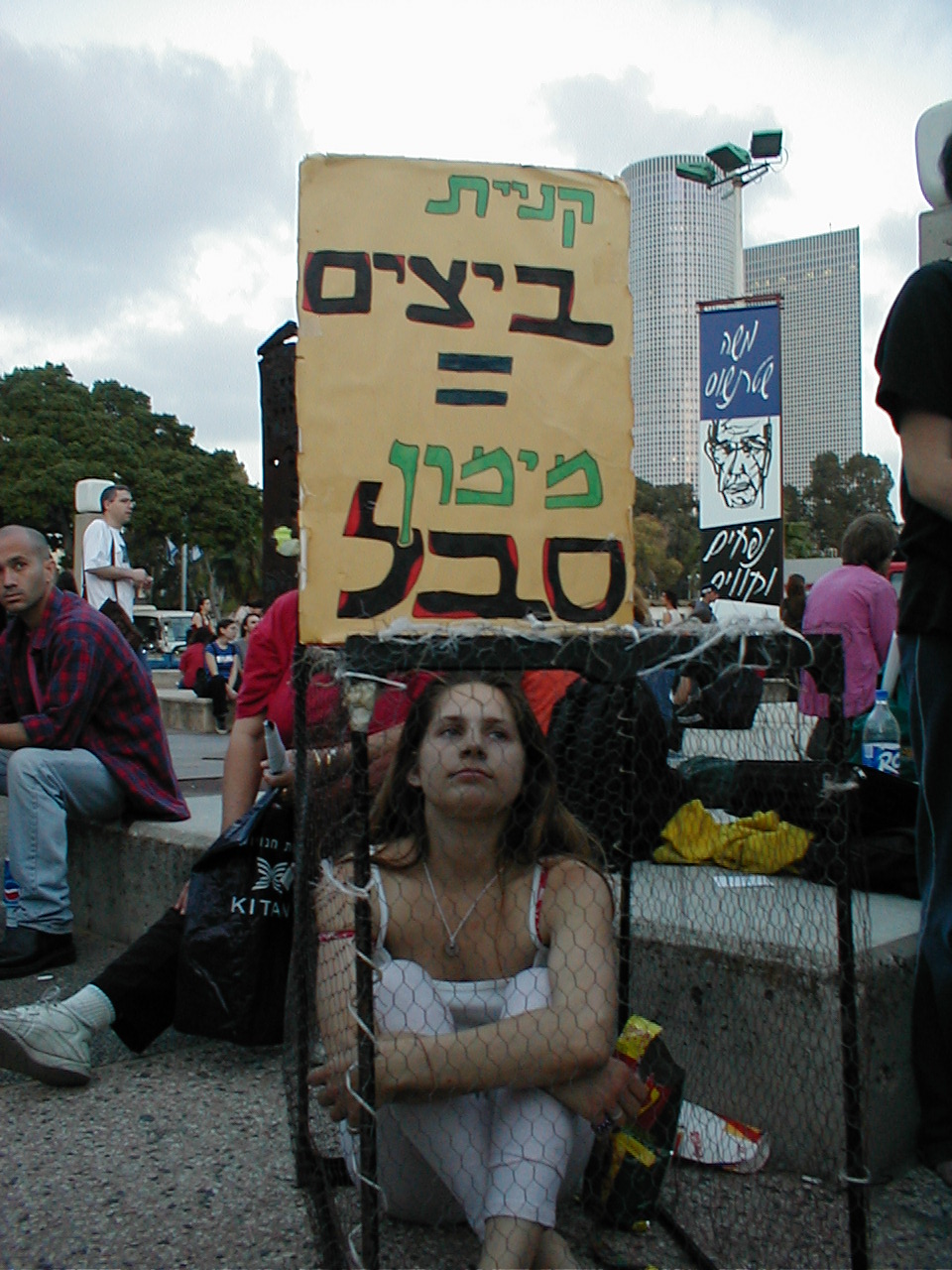 a woman sits behind a protest sign in an outdoor setting