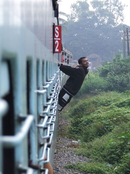 man hanging off the side of a train near grass and trees
