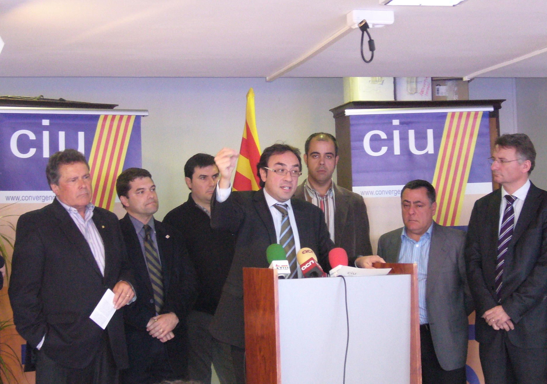 five men wearing ties are standing in front of a podium while others stand in the background