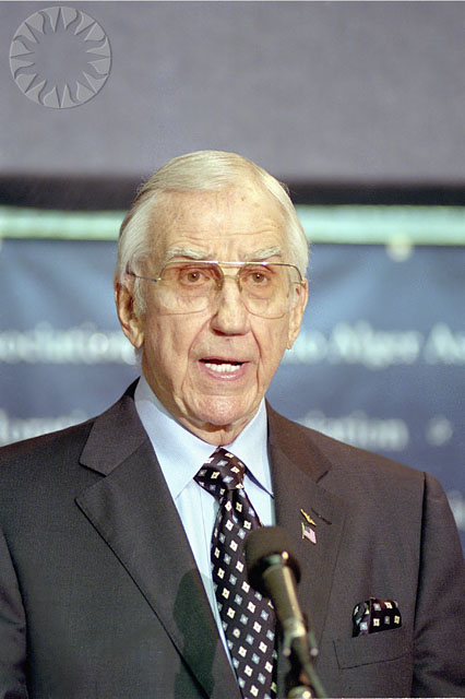 an older man in a business suit and tie talking into a microphone