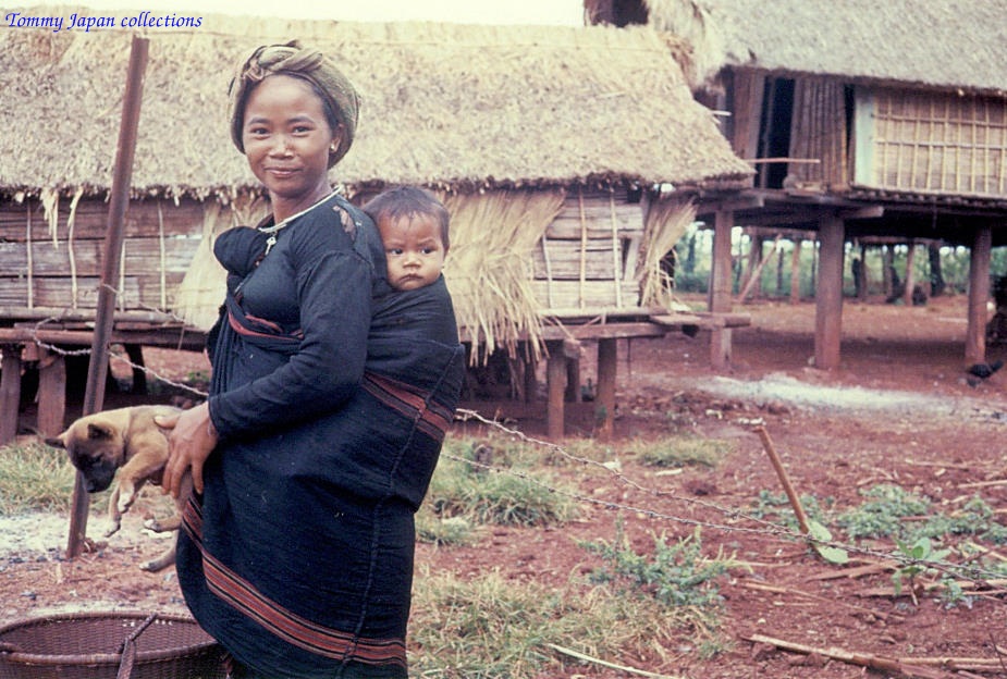 a woman carrying a child across a dirt ground near some huts