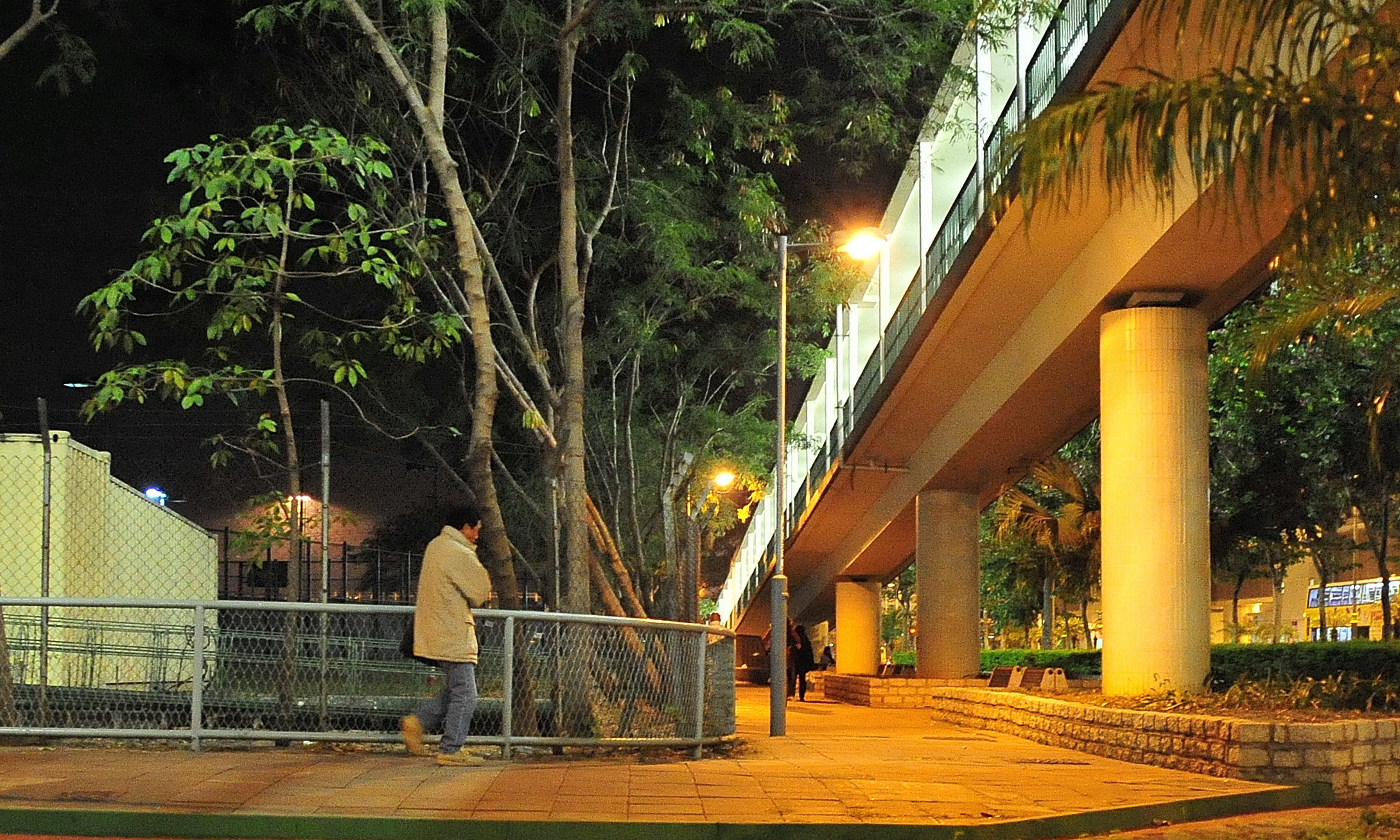 man in beige coat walking away from the fenced area under the bridge at night