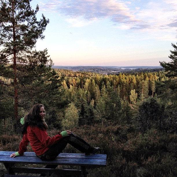 a woman wearing a red shirt is sitting on a wooden bench in the wilderness