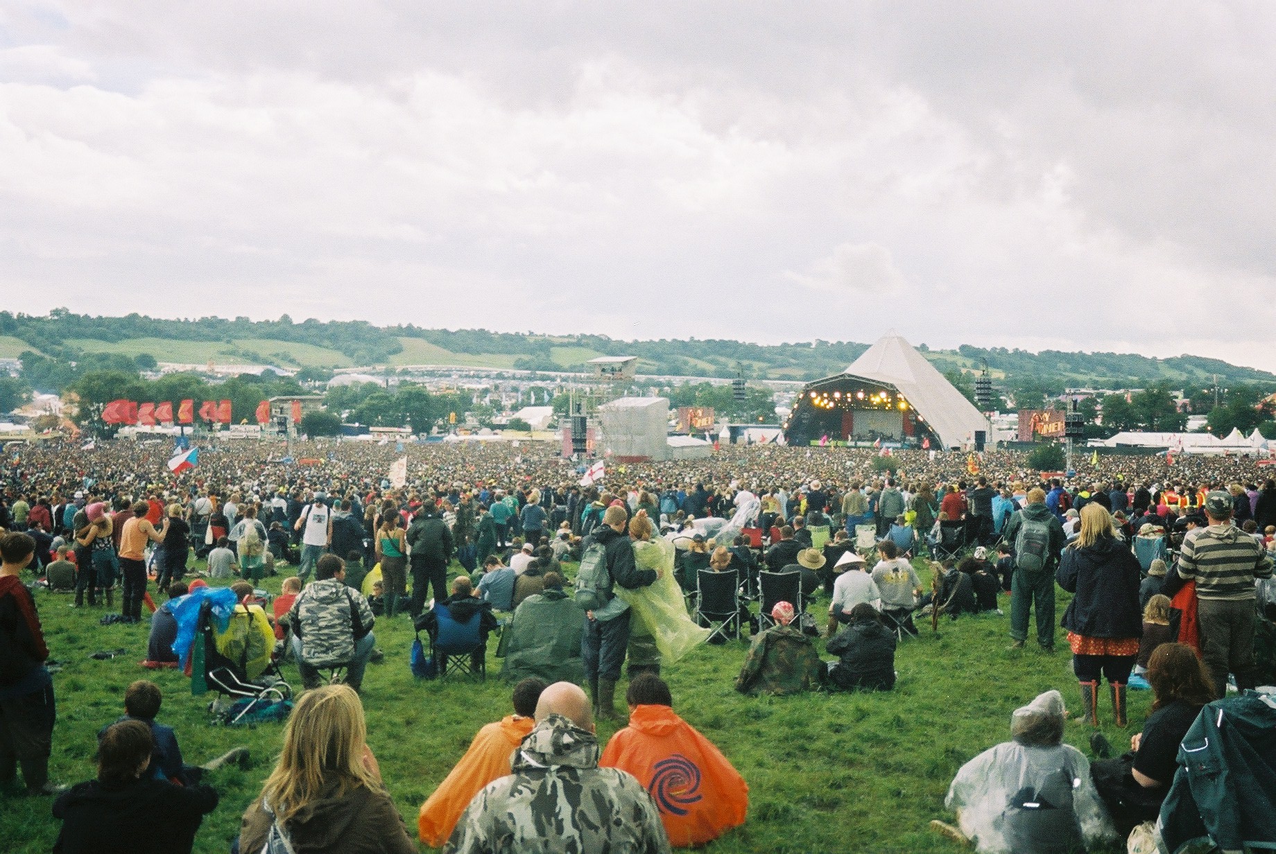 a crowd is at an outdoor concert, some with blankets and some with a laptop on their shoulders