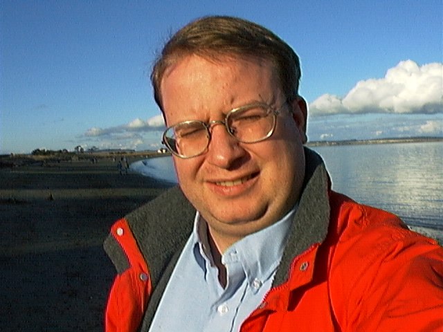 a man with glasses standing next to a body of water