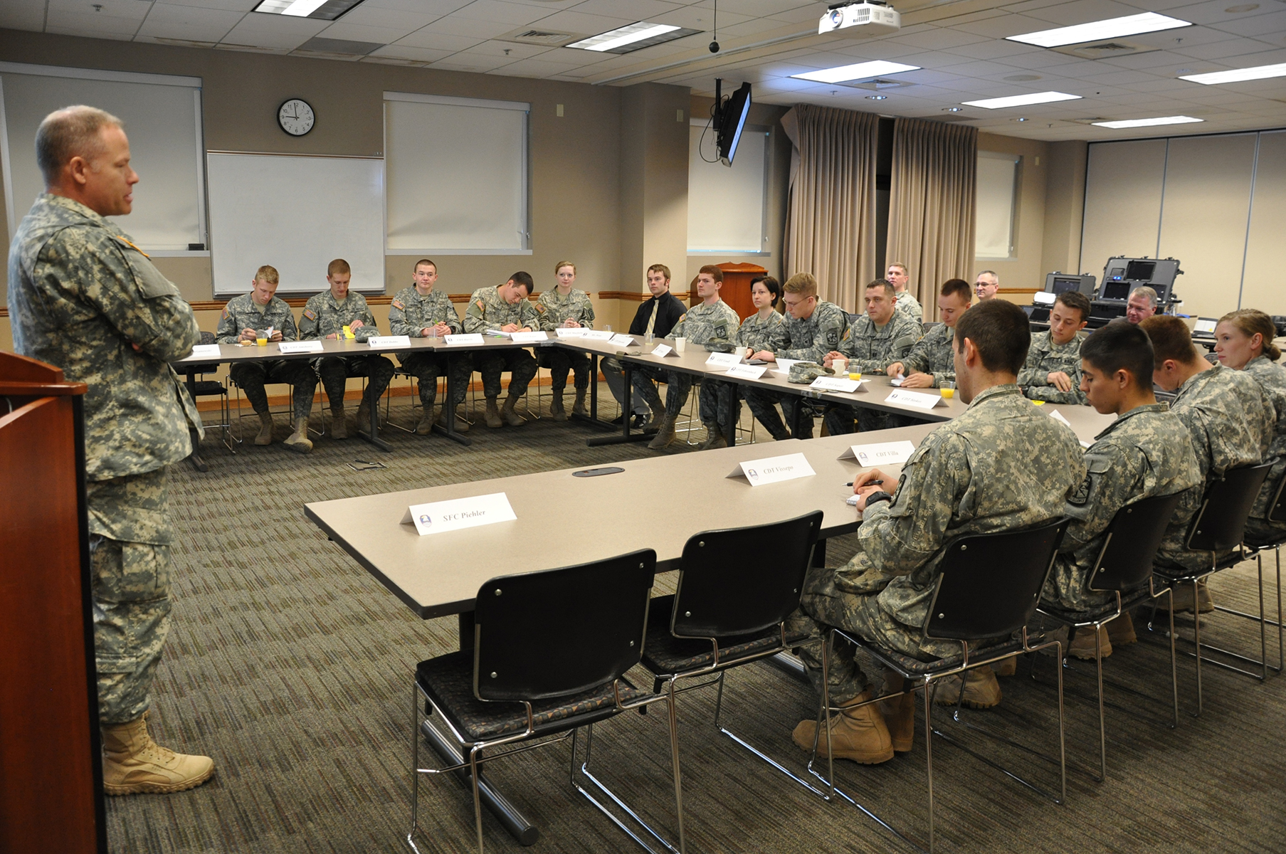 soldiers sit in a room with tables and chairs, facing an officer speaking