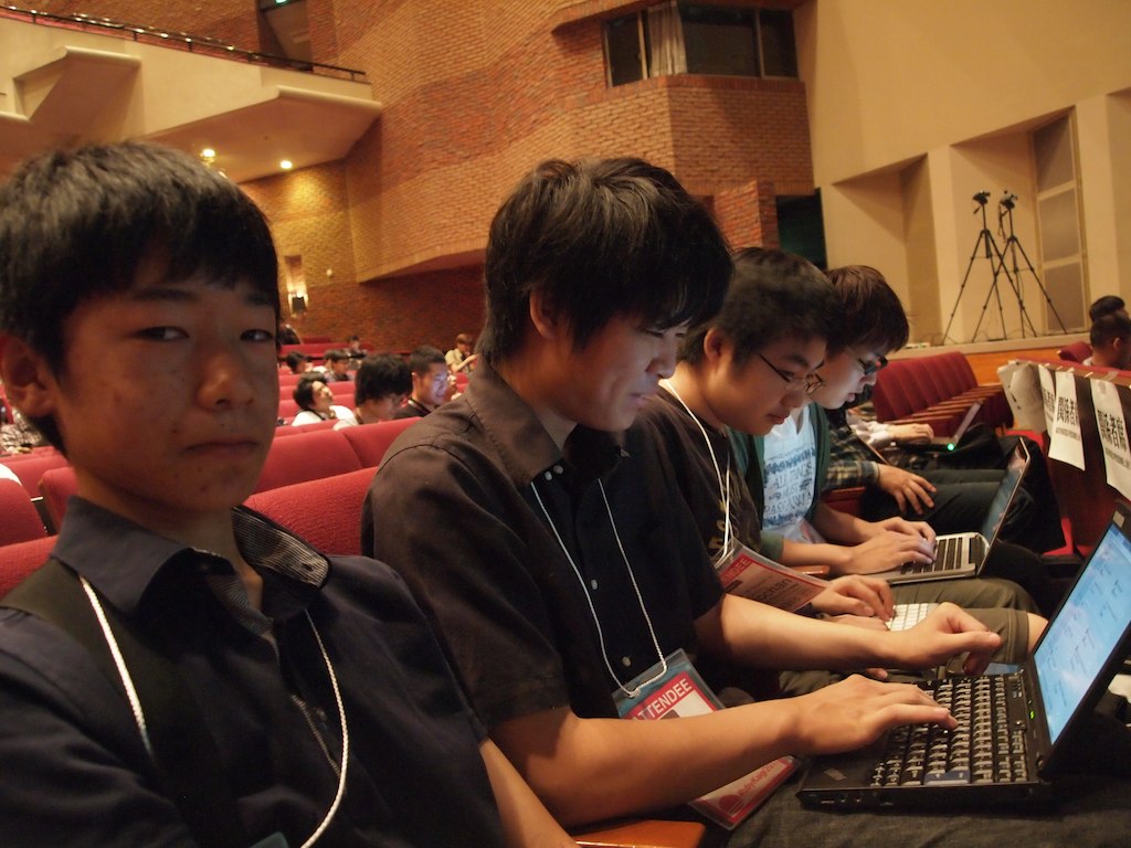 four boys sitting in auditorium using laptops