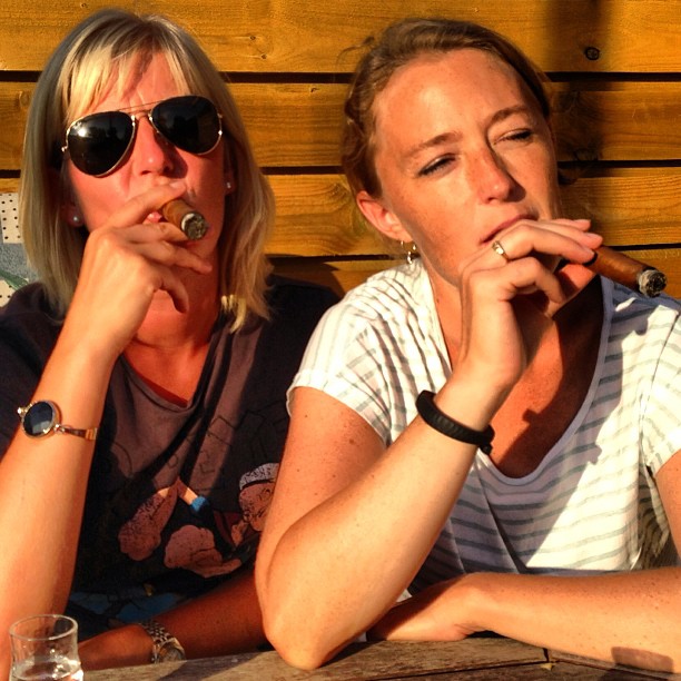 two people sitting on a wooden bench and smoking cigars