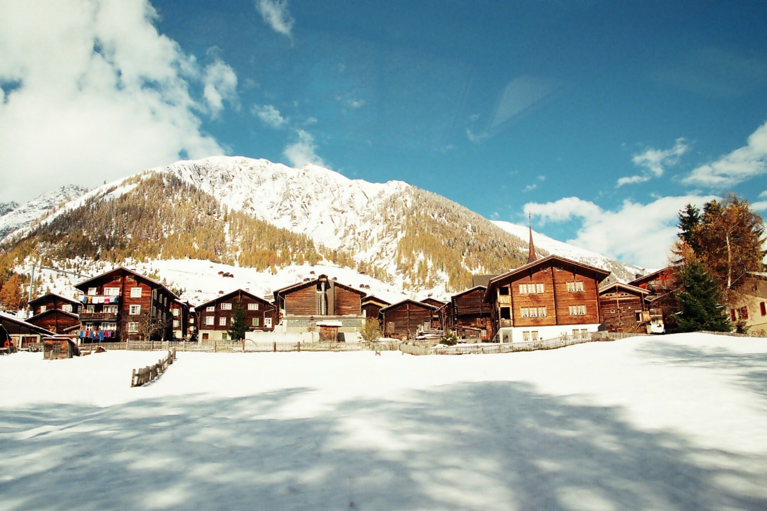 several wooden buildings in a snowy field with mountains in the background