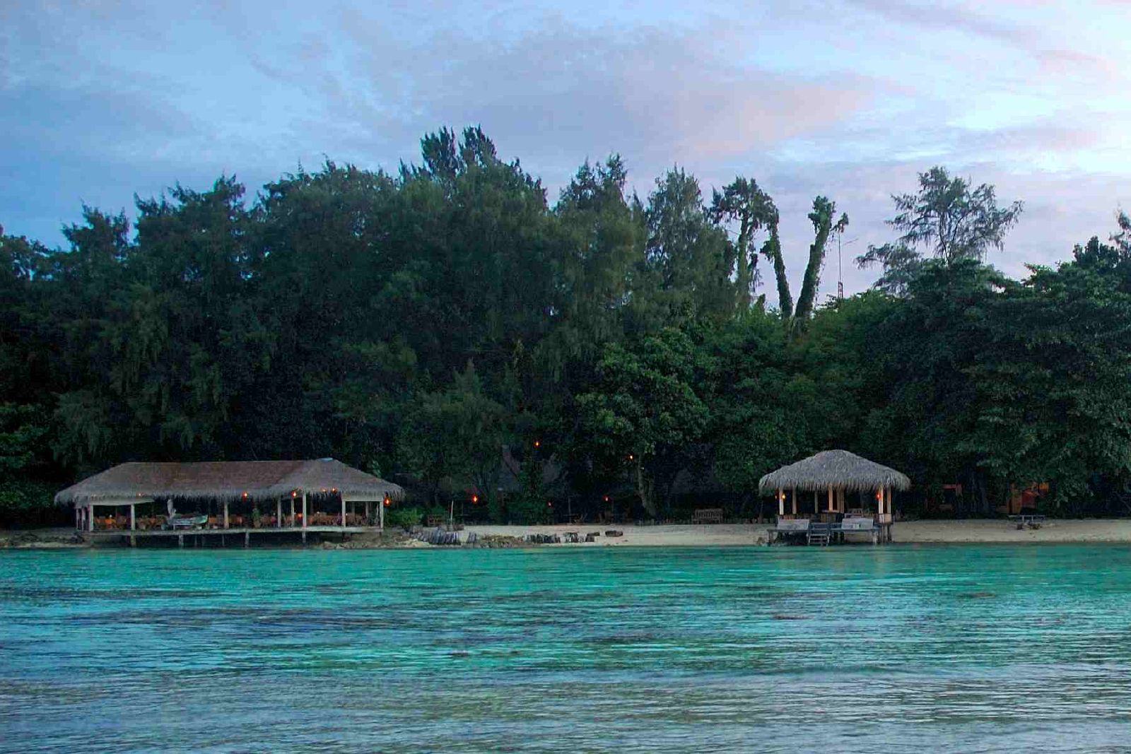 two huts on the beach are next to the trees