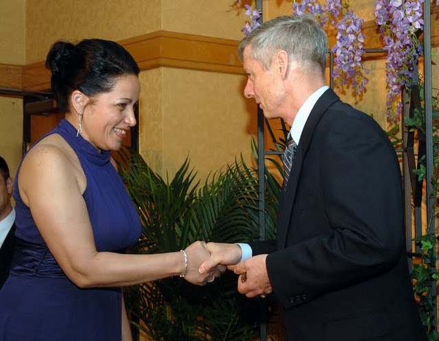 a smiling woman wearing a blue dress is shaking hands with an old man in front of an indoor plant