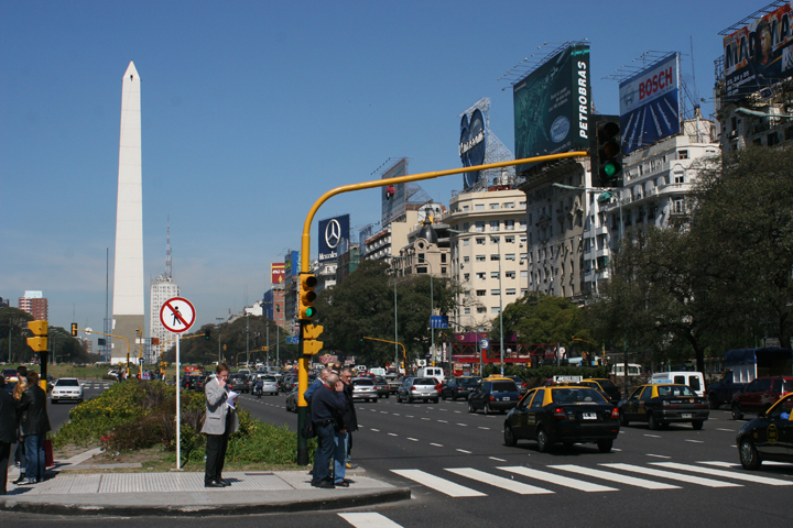 several people are standing at the edge of a busy city intersection
