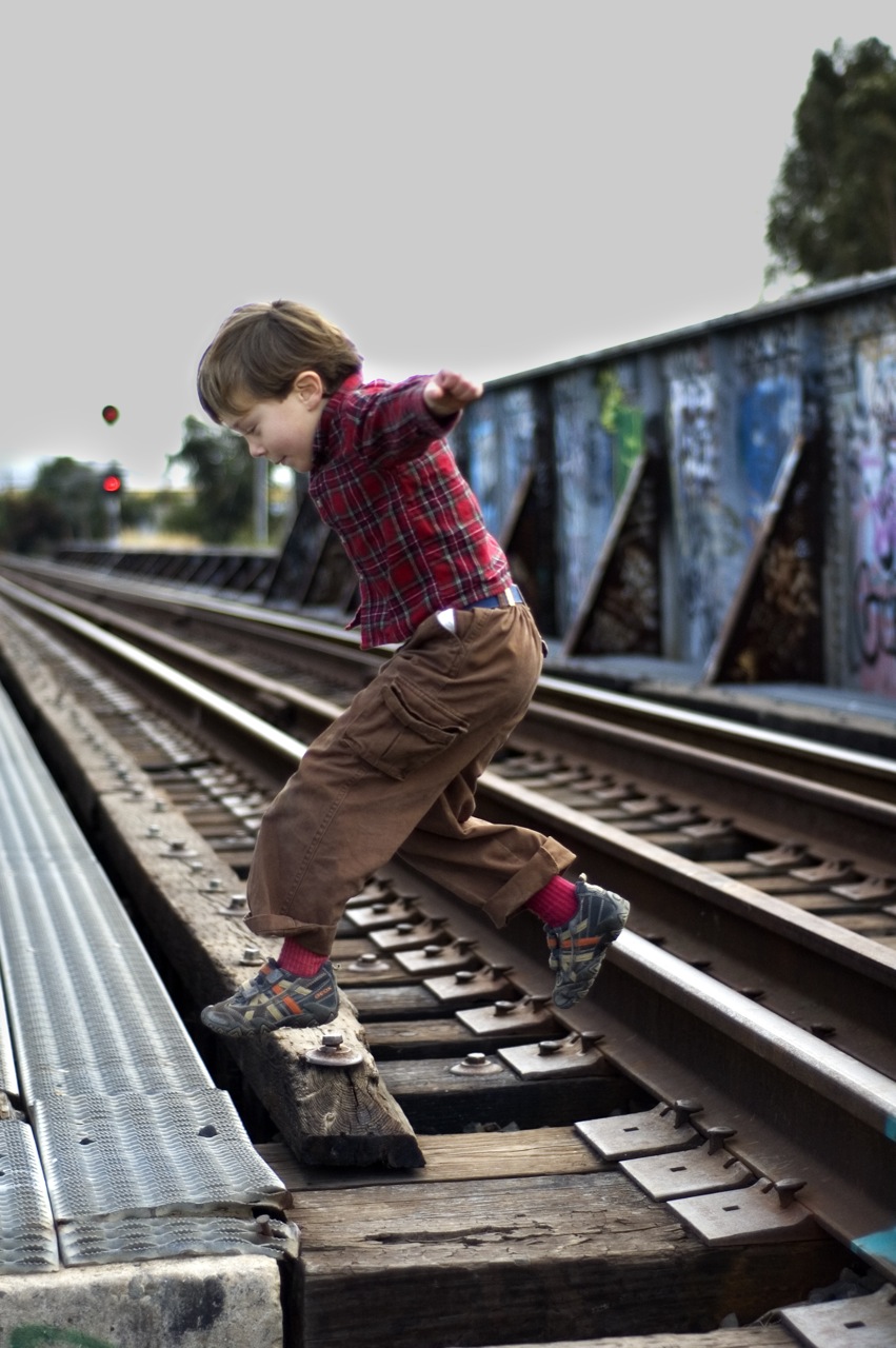 a boy wearing brown pants, red and white shirt and tan shoes skateboarding on railroad tracks
