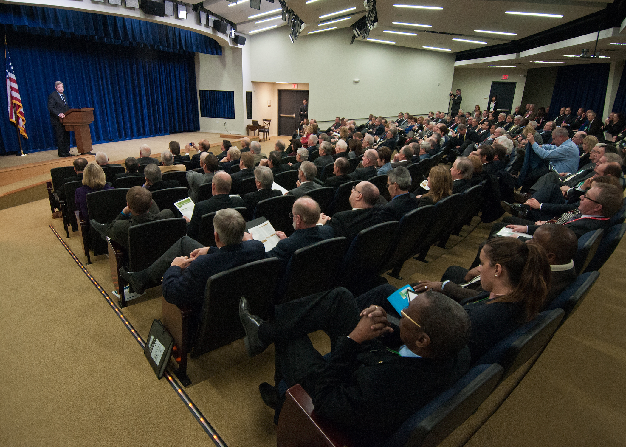 a lecture hall with two people sitting in chairs at the front of the room, watching soing while someone in the background stands in front of the audience