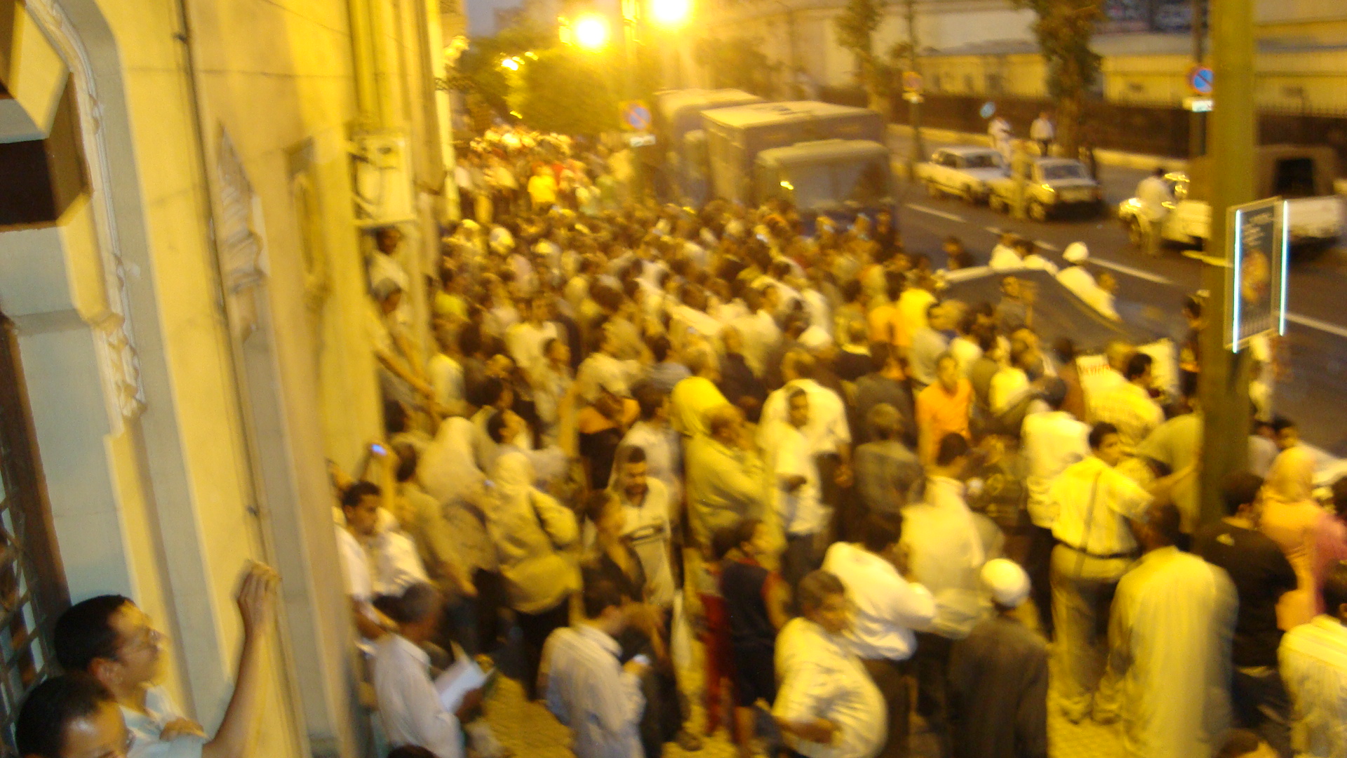 a crowd of people in white stands on the sidewalk near some parked cars