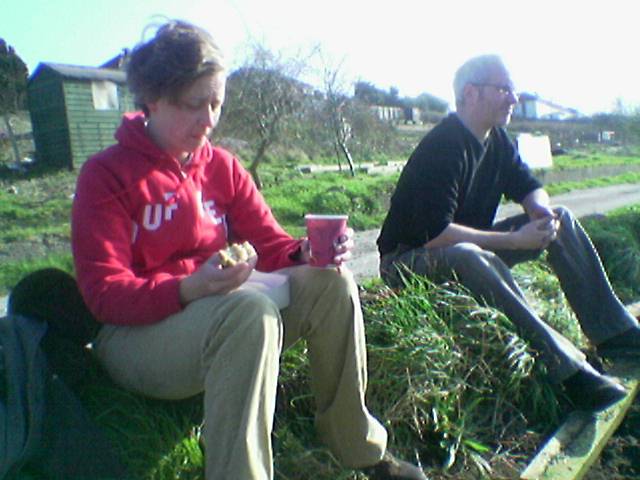 a man and woman sitting in the grass, one drinking coffee and the other looking on