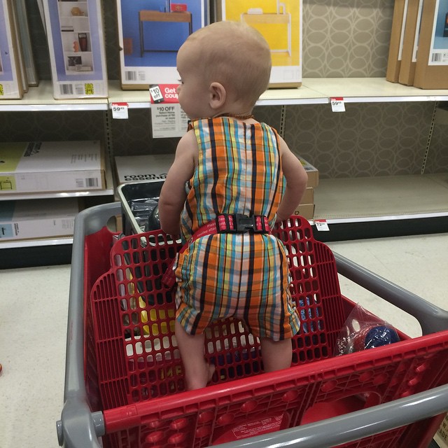 baby in red shopping cart with one foot on it in aisle