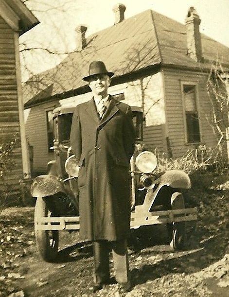 an old black and white po of a man wearing a suit and hat in front of a motor vehicle