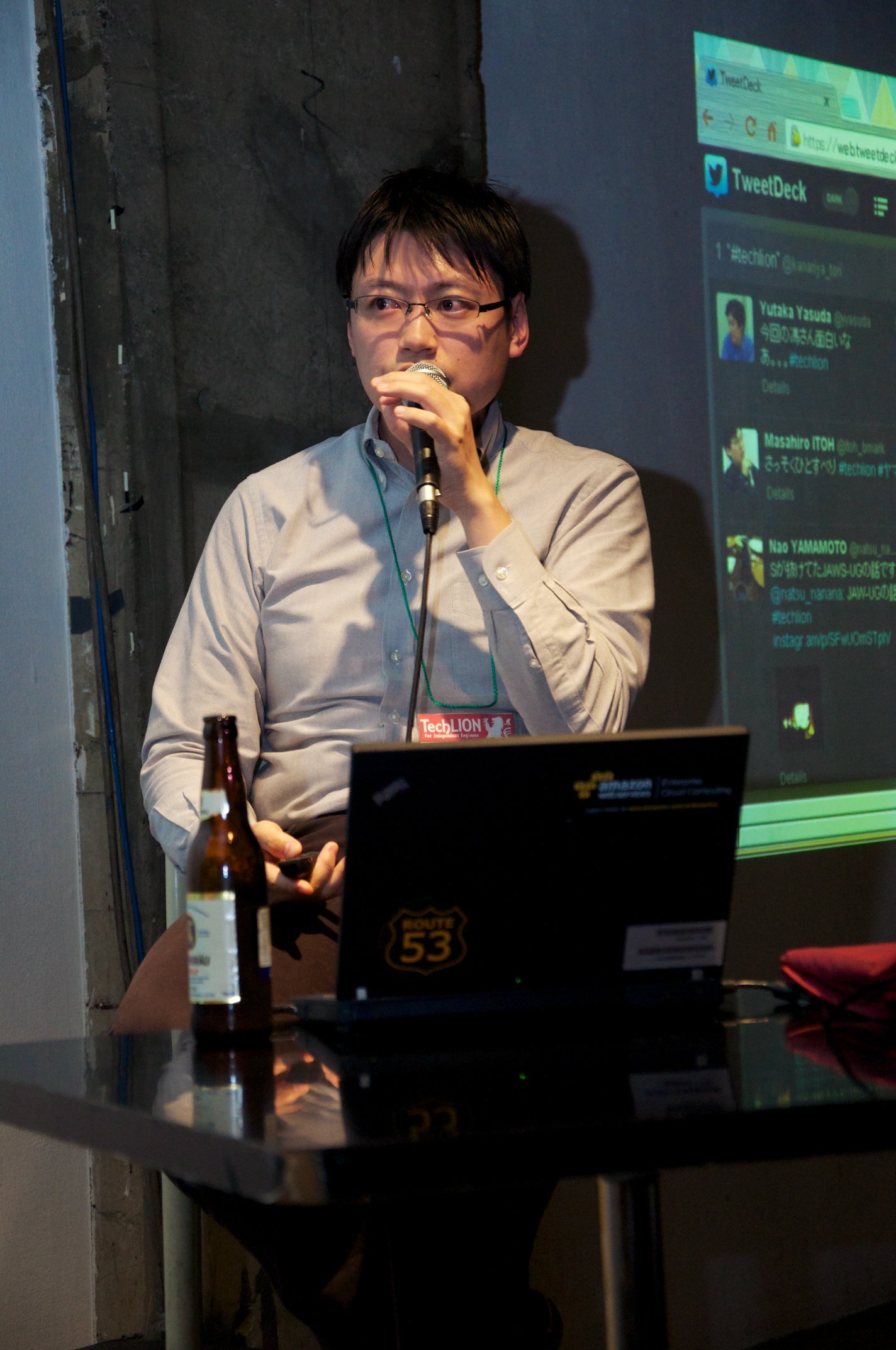 man in glasses, standing behind a desk with a laptop on it while talking into a microphone
