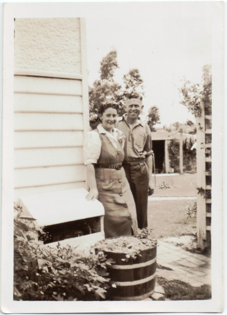 black and white pograph of a man and woman standing in front of a shed with a plant pot next to a wheel