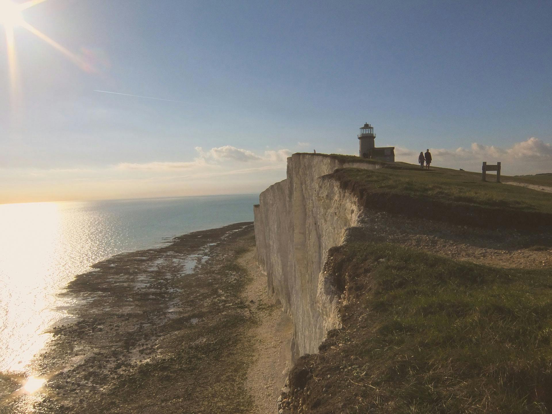 a couple of people standing on top of a cliff, by Rachel Reckitt, pexels contest winner, chalk cliffs above, light house, panoramic, late summer evening