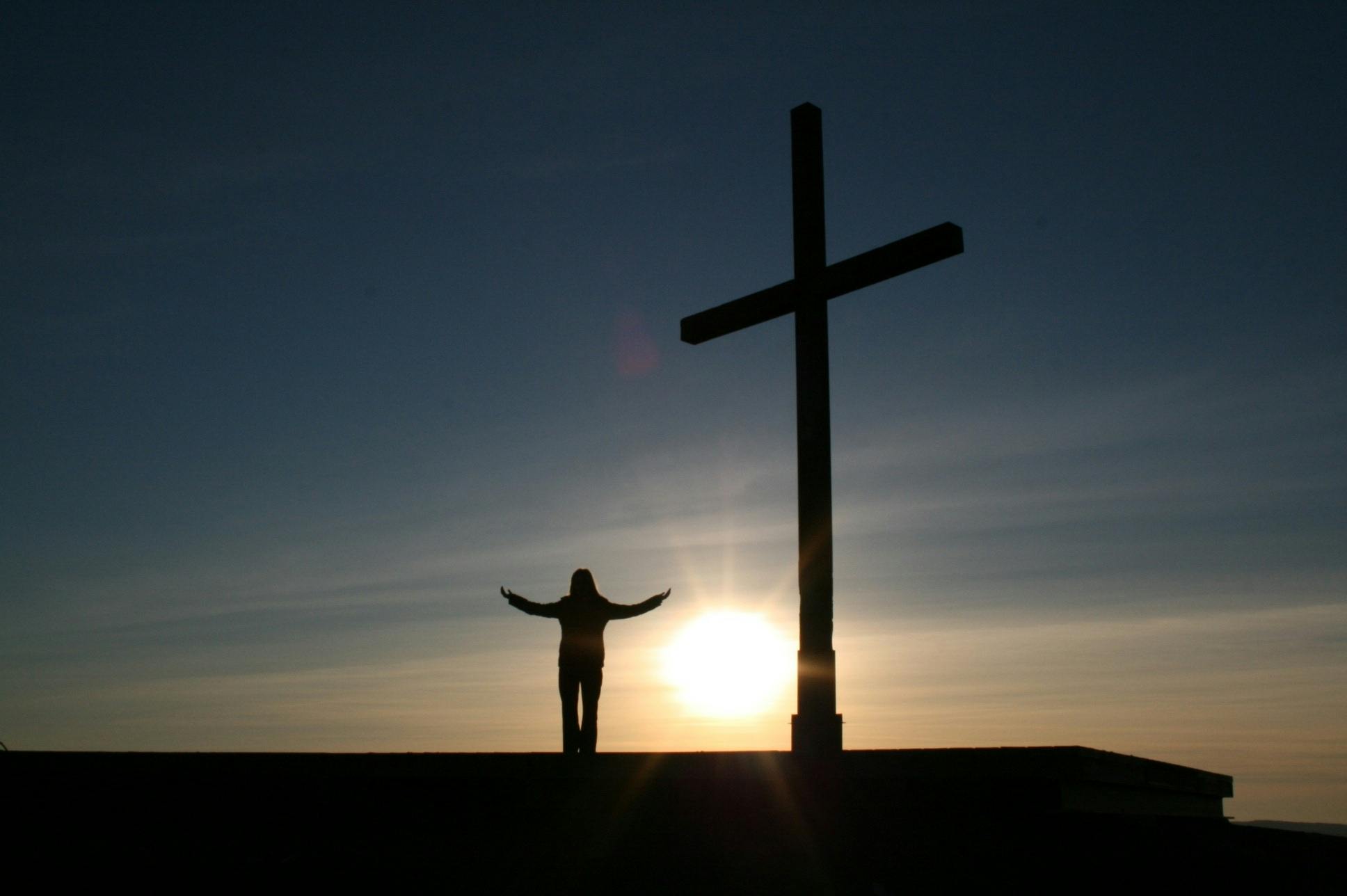 a person standing in front of a cross at sunset, with arms up, dimly - lit, image, on a sunny day