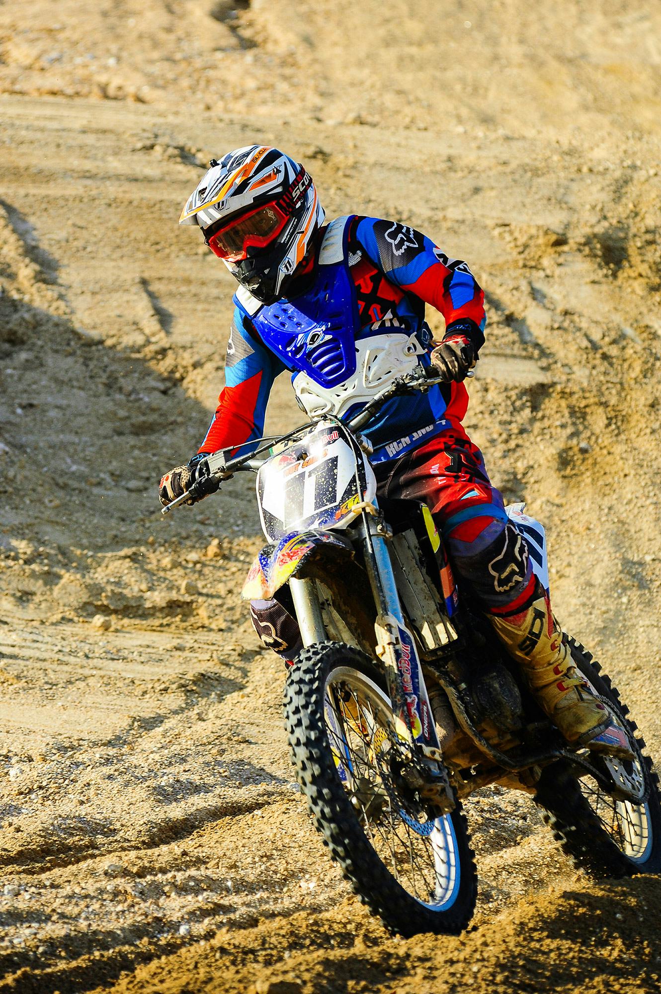 a man riding on the back of a dirt bike, profile image, zoomed in, red blue and gold color scheme, sports clothing