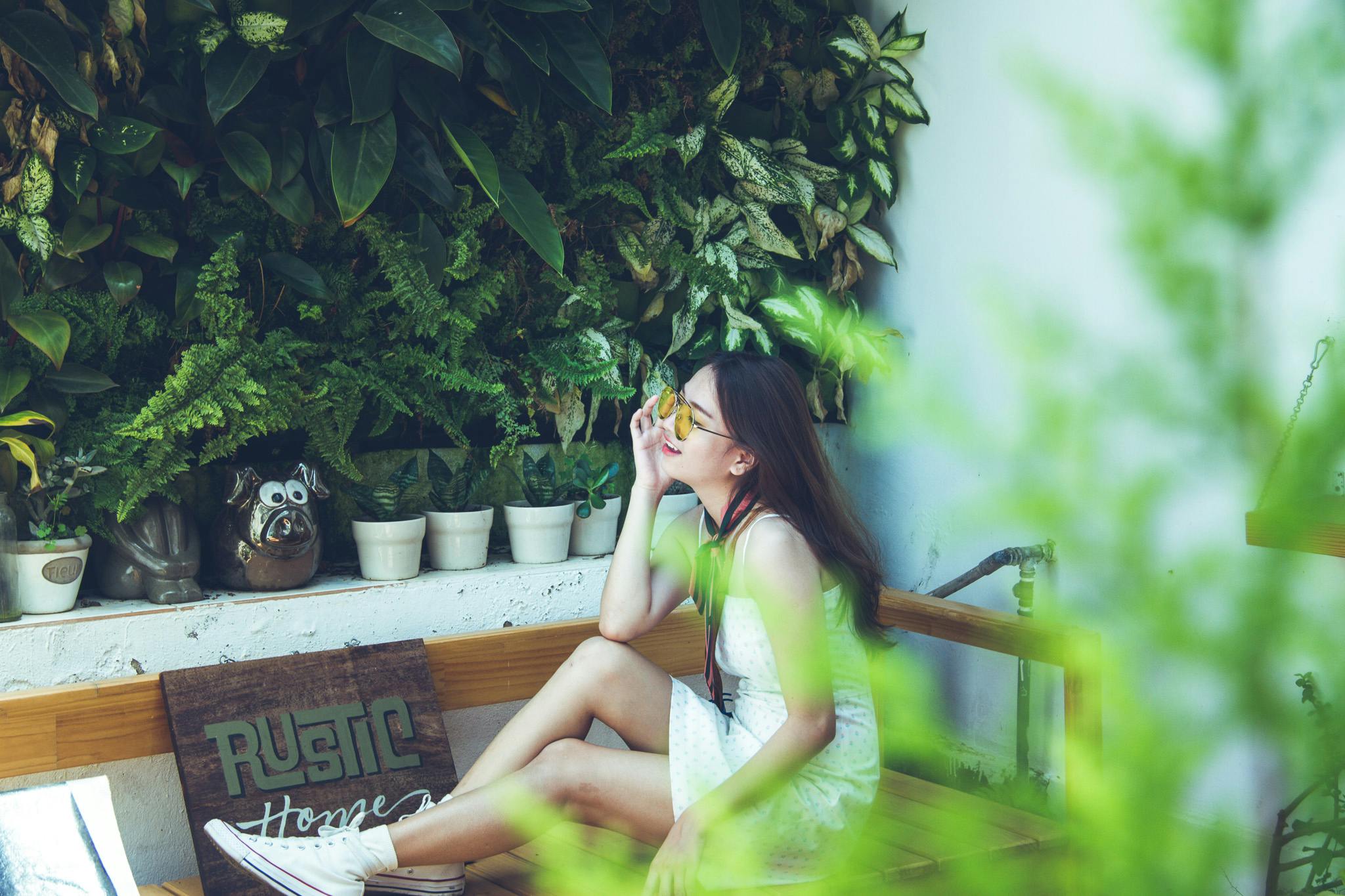 a woman sitting on top of a wooden bench, inspired by Elsa Bleda, trending on pexels, verdant plant wall, girl with glasses, teenager hangout spot, yellow and greens