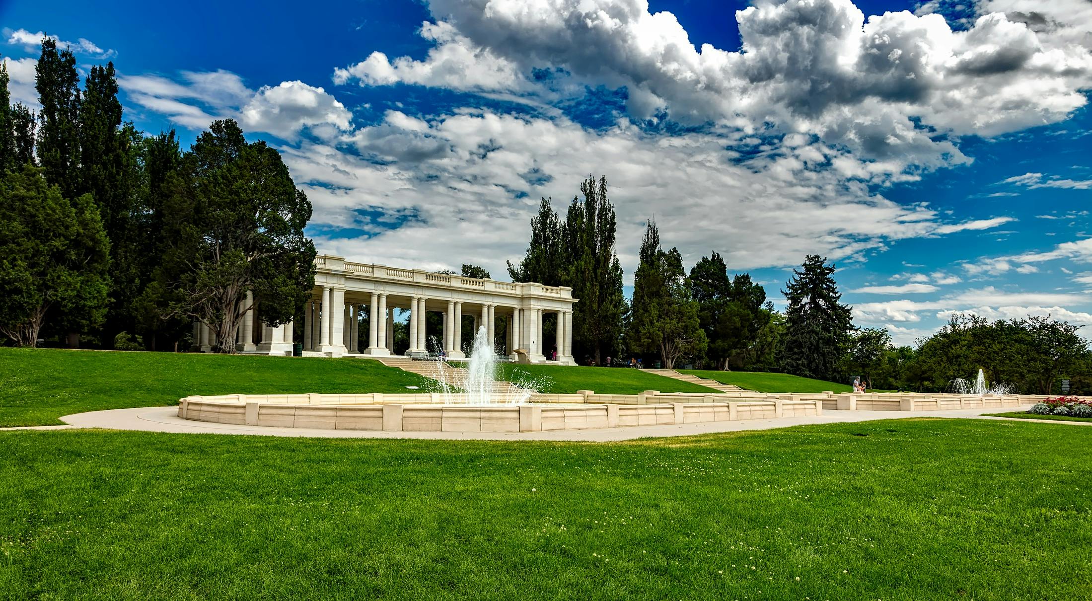 a fountain sitting on top of a lush green field, a marble sculpture, colonnade, colorado, in a city with a rich history, ceremonial clouds