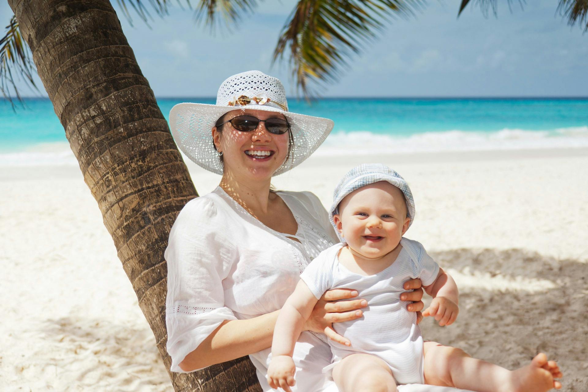 a woman holding a baby under a palm tree, white suit and hat, square, on a sunny beach, seraphine