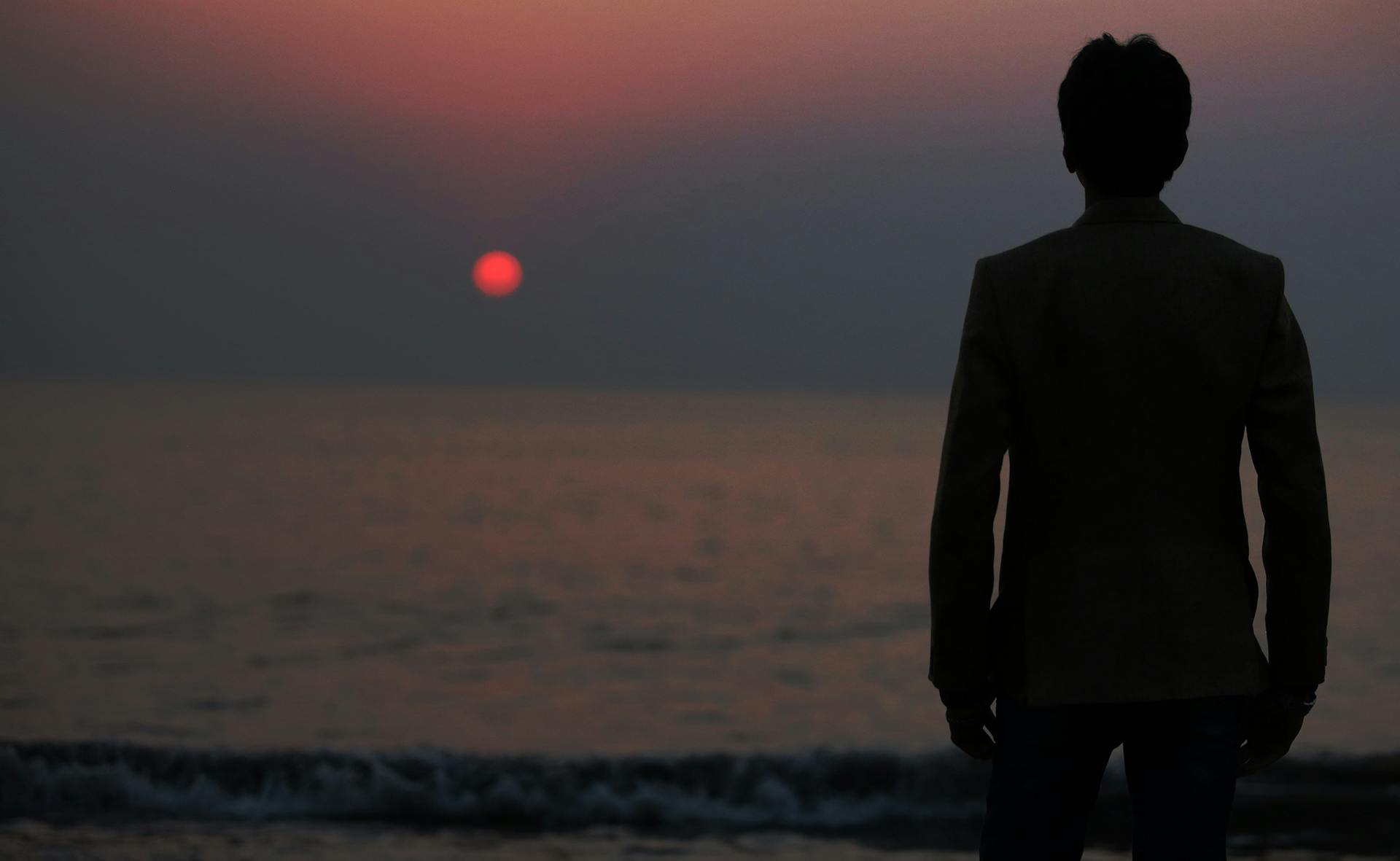 a man standing on top of a beach next to the ocean, an album cover, pexels contest winner, romanticism, at gentle dawn red light, distant thoughtful look, still from a terence malik film, the sun at their back