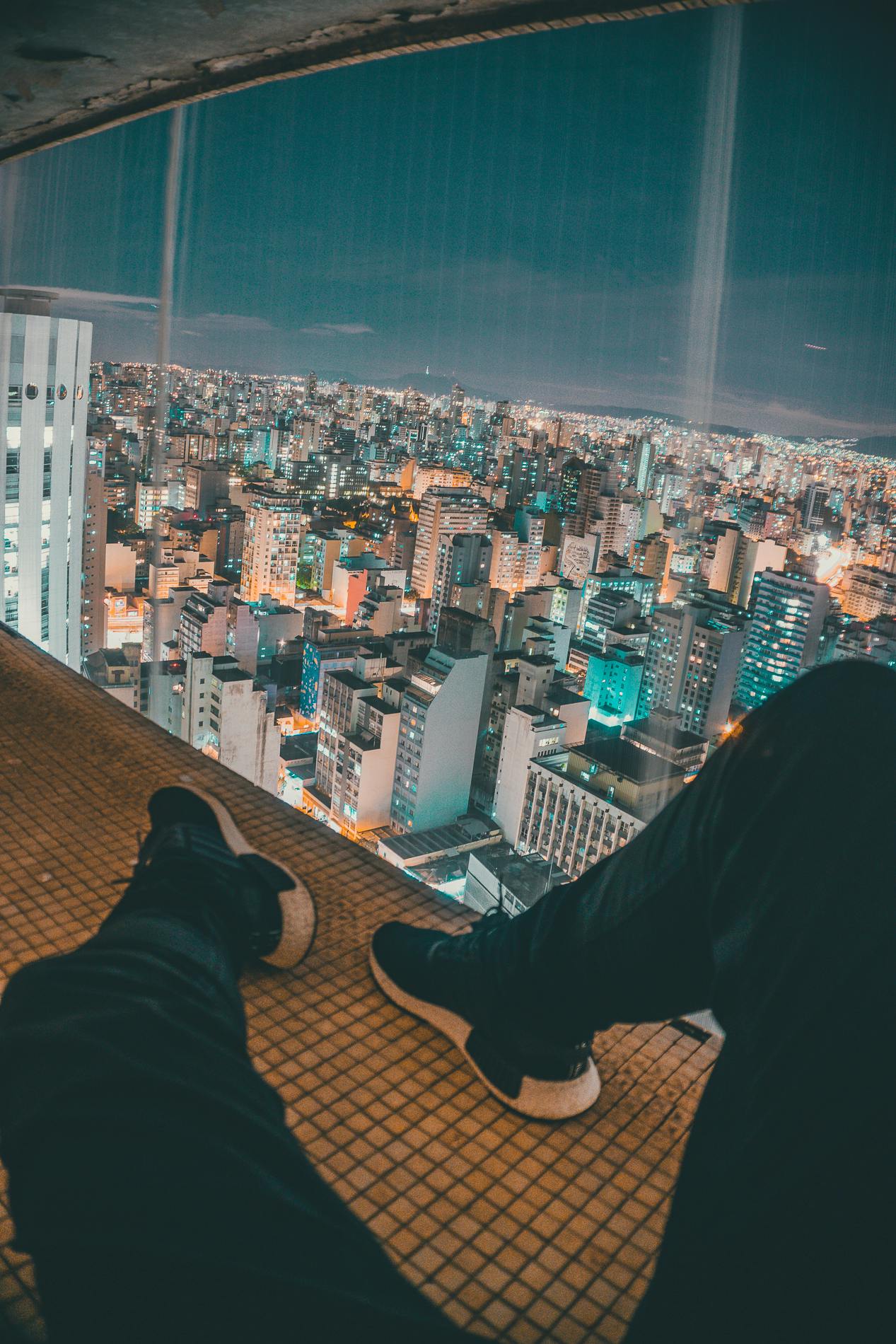 a person sitting on a ledge overlooking a city, by Niko Henrichon, pexels contest winner, futuristic sao paulo, sitting on the floor, high soles, high lights