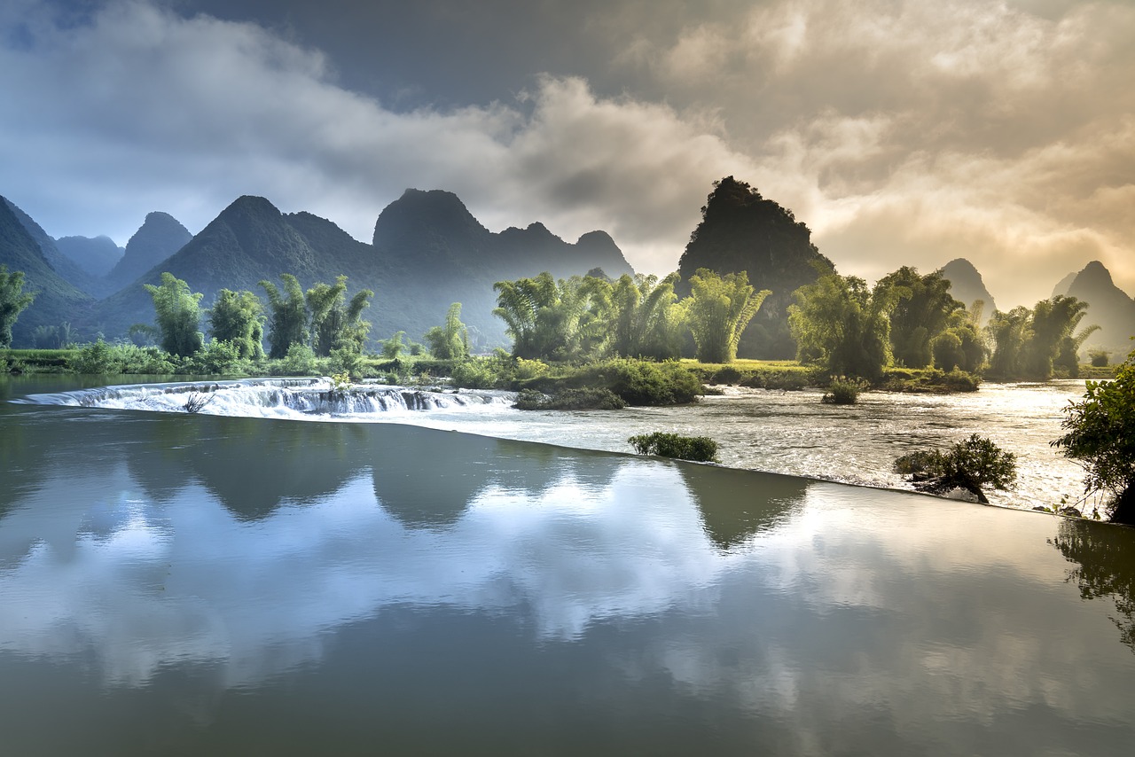 a body of water with trees and mountains in the background, a picture, by Etienne Delessert, shutterstock, vietnam, flowing water, springtime morning, water reflecting suns light