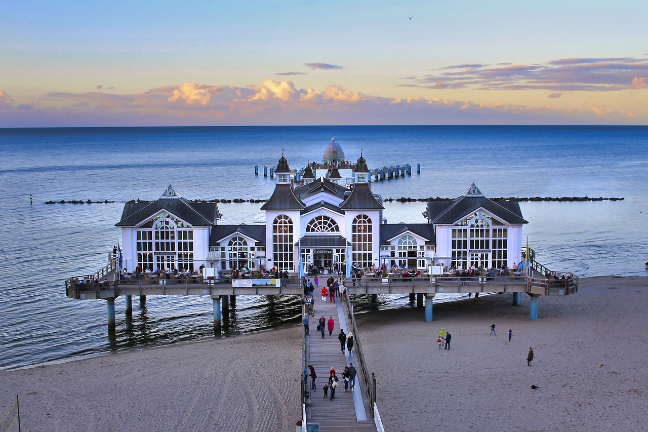 a group of people walking on a beach next to a pier, by Emanuel Büchel, shutterstock, seaside victorian building, in the evening, above view, lower saxony