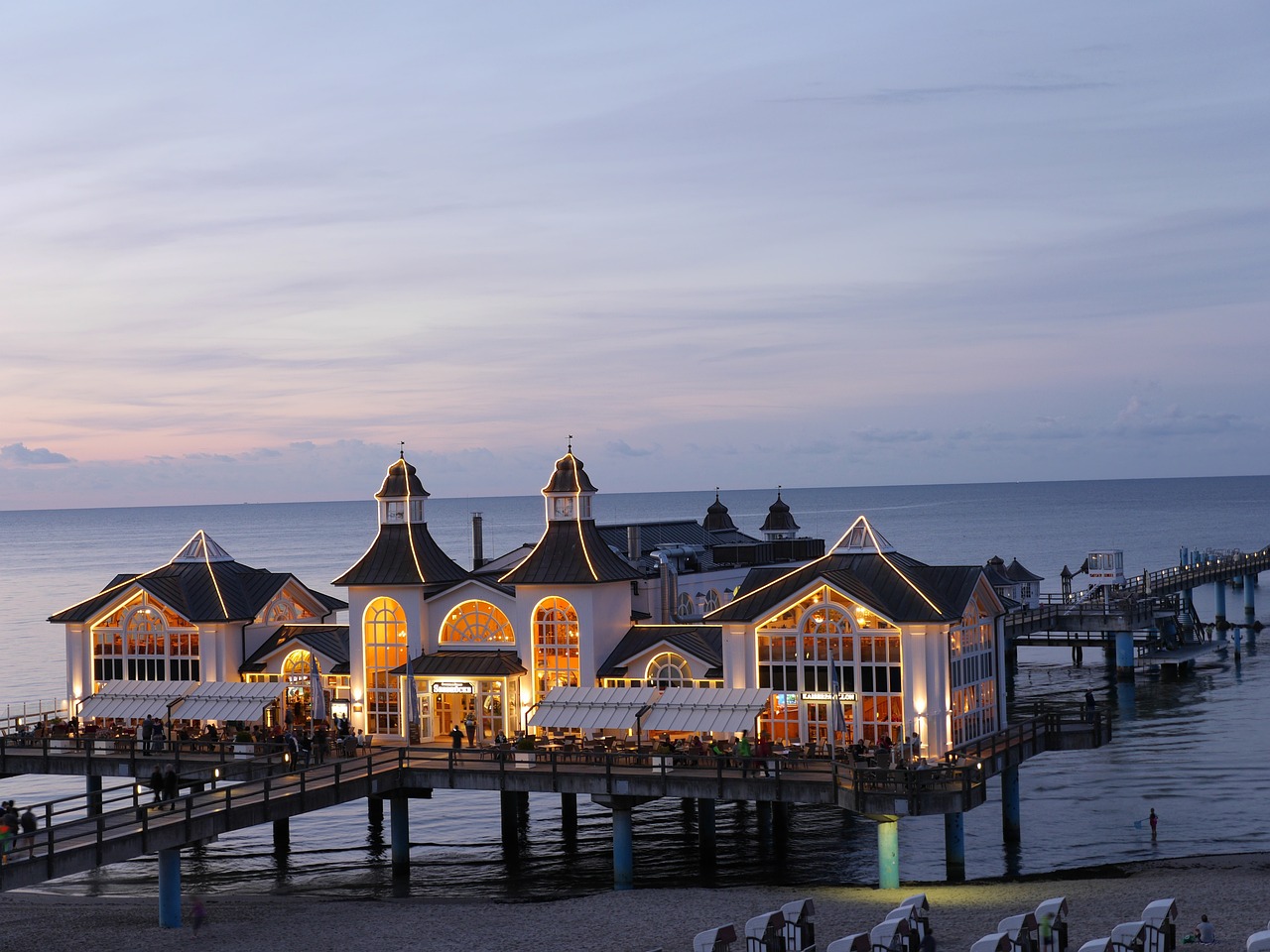 a group of people standing on top of a pier next to a body of water, by Juergen von Huendeberg, pixabay, art nouveau, restaurant in background, sitting on the beach at night, seaside victorian building, lower saxony