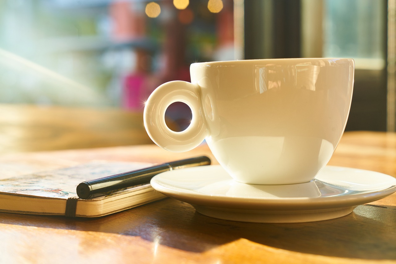 a coffee cup sitting on top of a saucer next to a book, by Karl Buesgen, in a bright cafe, istock, morning detail, iphone