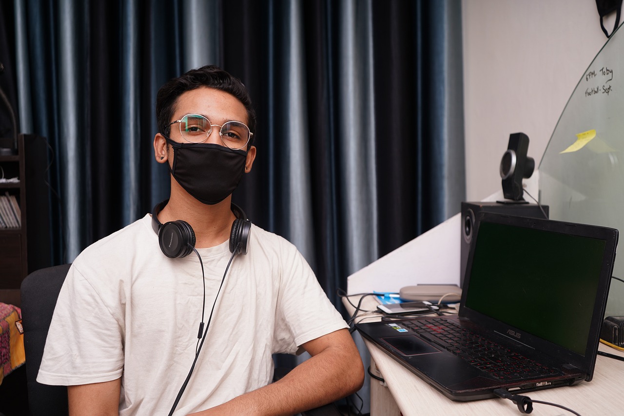 a man sitting in front of a laptop wearing a face mask, realism, around 1 9 years old, posing for the camera, indoor lab scene, wearing black headphones