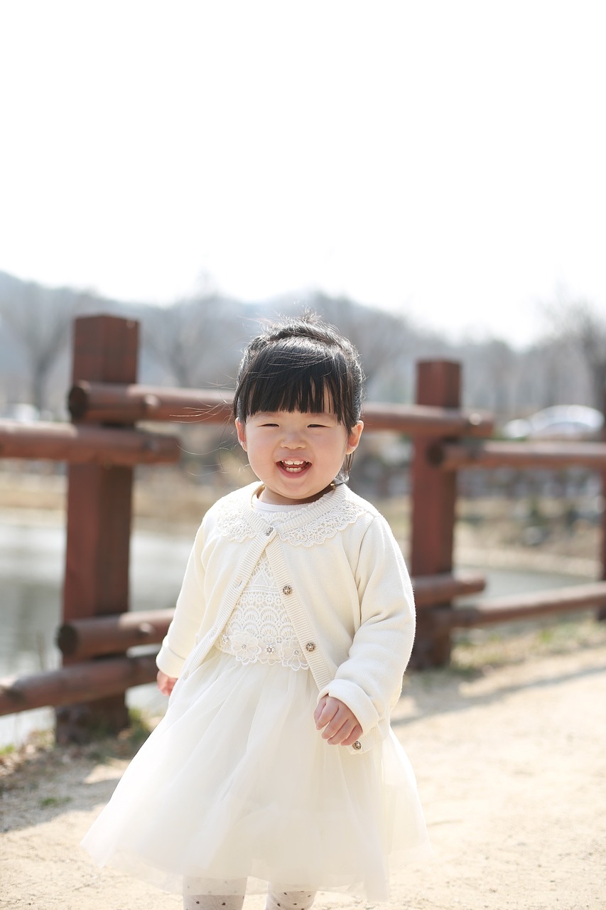 a little girl in a white dress standing in front of a fence, by Simon Gaon, pexels, mingei, wan adorable korean face, [[[[grinning evily]]]], walking at the park, 4 k ]