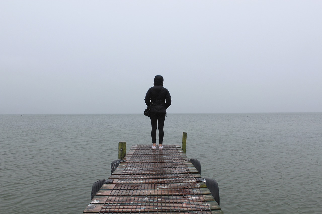 a person standing on a dock looking out at the water, minimalism, girl in raincoat, dangerous depressing atmosphere, wide shot photo